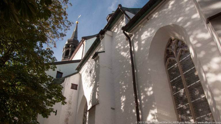 Low-angle view of a white historic church with a tall pointed Gothic window and dark spire rising behind the roof.