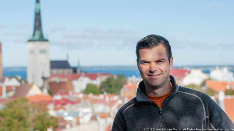 Adult posing at an elevated Tallinn viewpoint with red Old Town roofs and a pale church tower blurred behind.