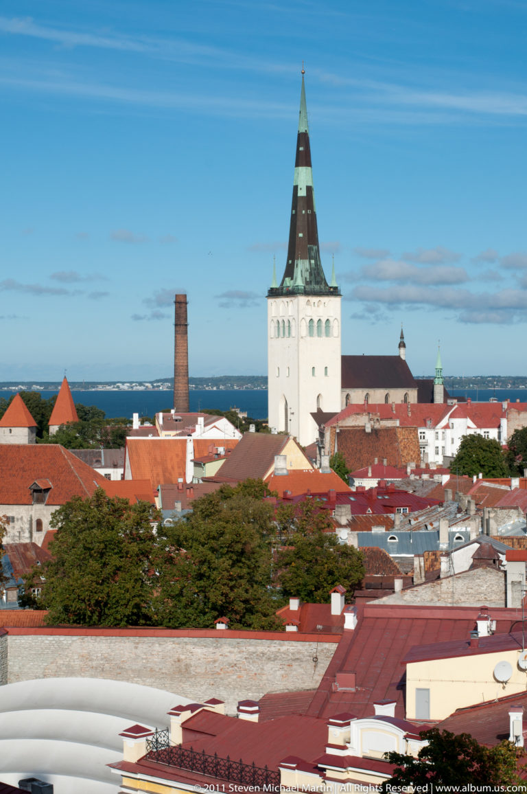White square church tower with a dark tapering spire rising above Tallinn’s red rooftops and distant blue horizon.