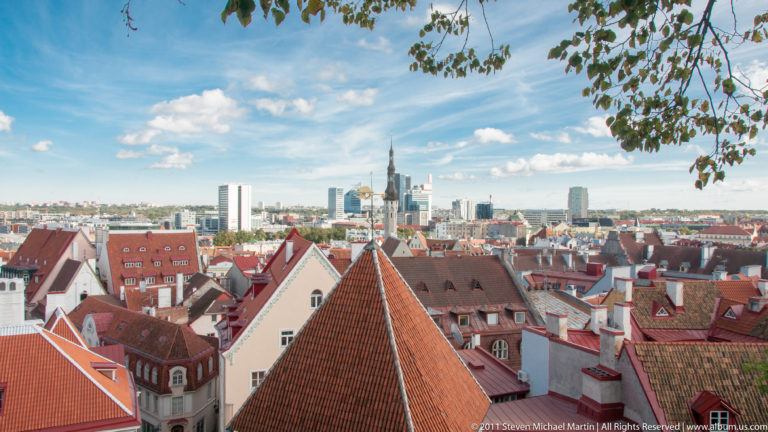Elevated Tallinn cityscape with steep Old Town roofs in the foreground and modern high-rise buildings on the horizon.