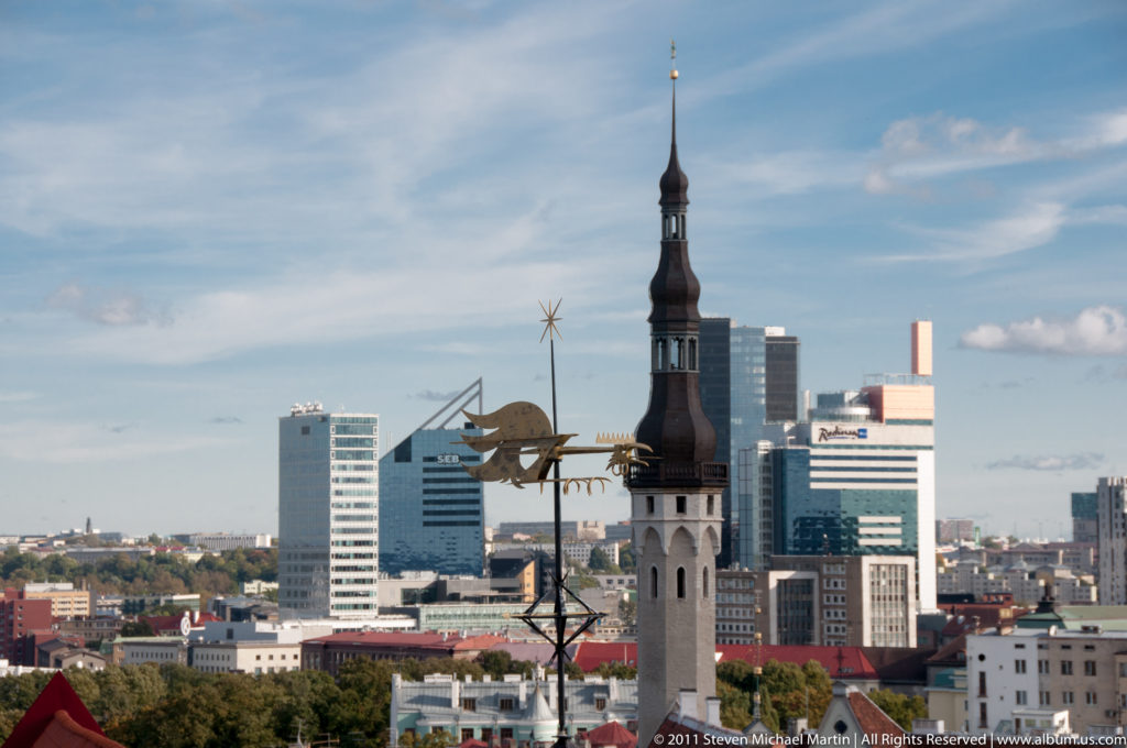 Historic church tower, foreground weather vane, and modern high-rises seen across Tallinn.