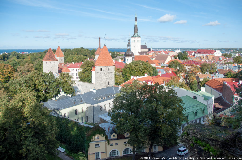 Elevated view across Tallinn Old Town with red-tiled roofs, medieval wall towers, trees, and a tall white church spire.