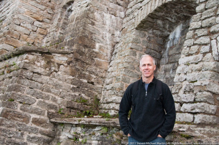 Adult standing before a deep arched recess in rough medieval stone masonry, showing the scale of the historic wall.