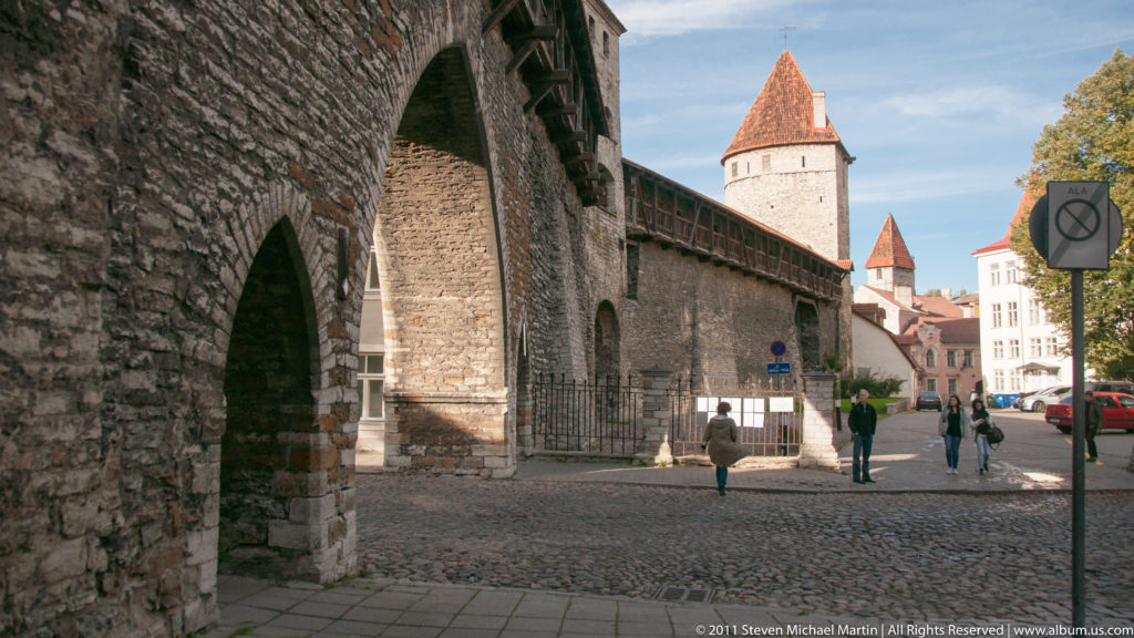 Street-level view of Tallinn’s medieval city wall with arched opening, timber wall-walk, and red-roofed defensive towers.