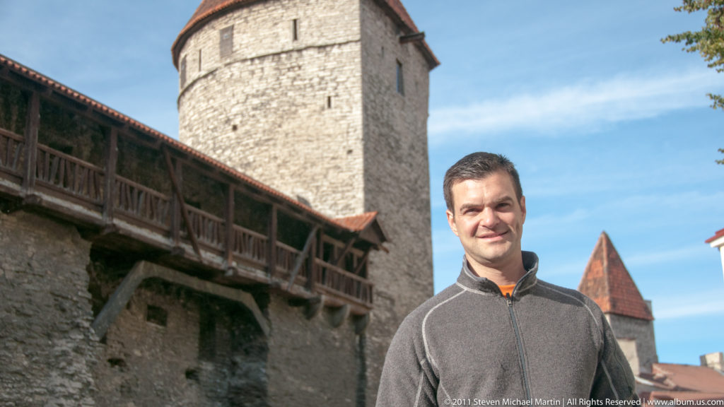 Adult standing beside Tallinn’s medieval stone wall and covered timber wall-walk, with defensive towers rising behind.
