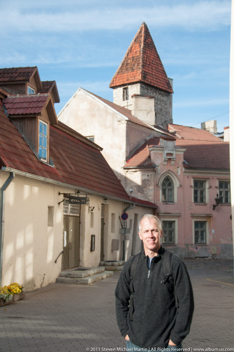 Adult standing in a paved Tallinn Old Town space with low historic buildings and a square medieval tower behind.