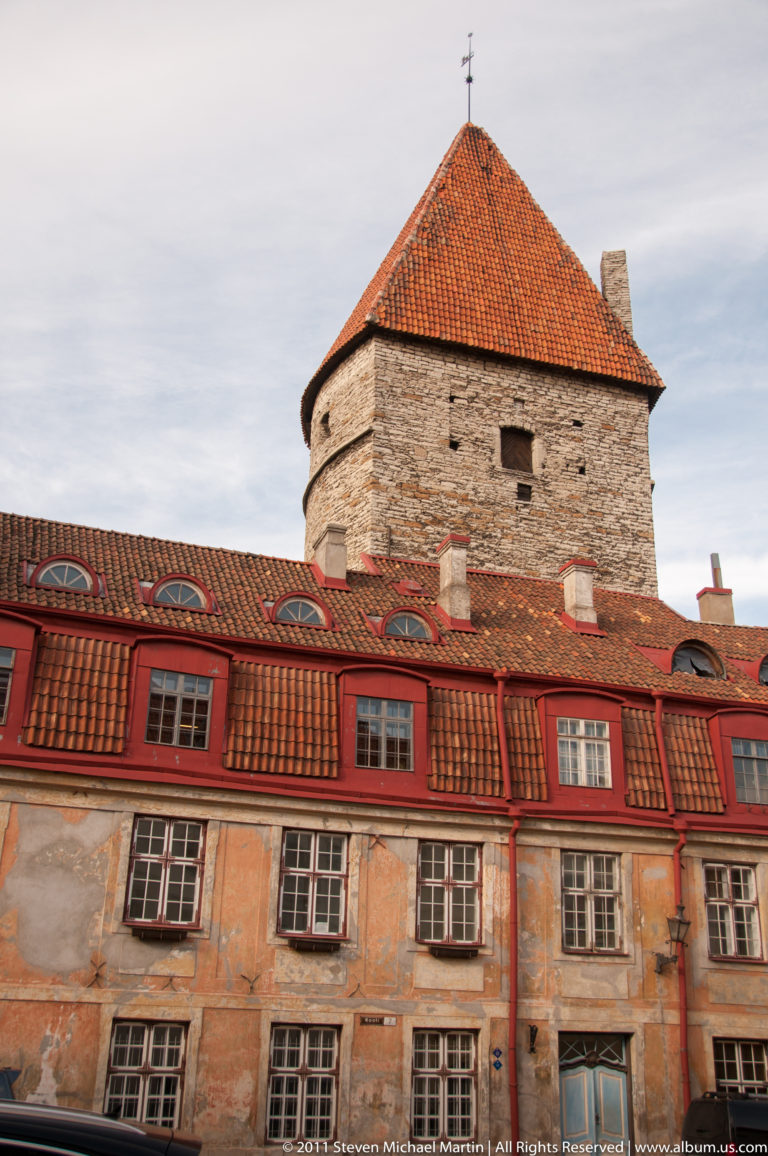 Large round medieval stone tower with red-tiled roof rising behind a weathered ochre building with rows of windows.