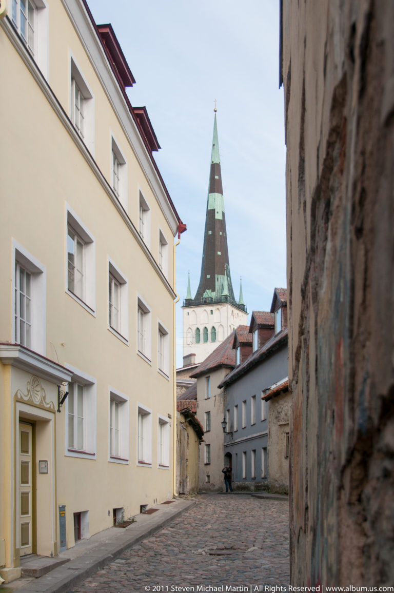Narrow Tallinn Old Town lane between pale historic buildings, with a tall green-capped church spire visible at the far end.