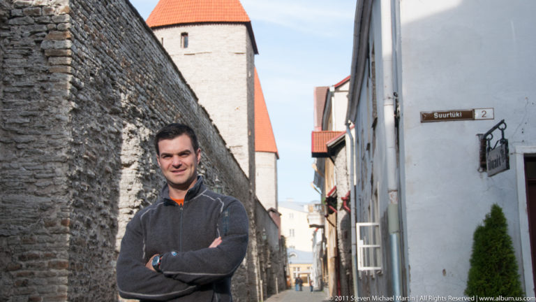 Adult standing beside Tallinn’s medieval stone wall on Suurtüki street, with a defensive tower and red-tiled roof behind.