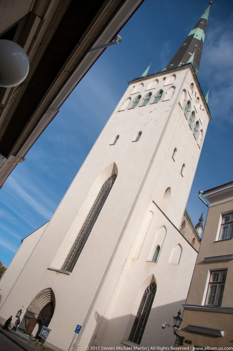 Low-angle view of a tall white Gothic church with pointed windows and steep spire rising above a narrow Tallinn Old Town street.
