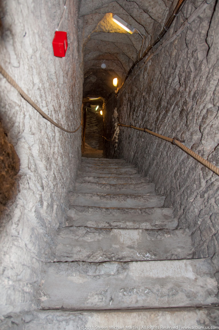 Steep stone stairway in a narrow rough-walled passage with handrails and small electric lights overhead.