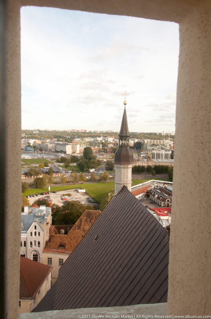 View through a rectangular masonry opening over Tallinn rooftops, a small tower roof, and the distant cityscape.