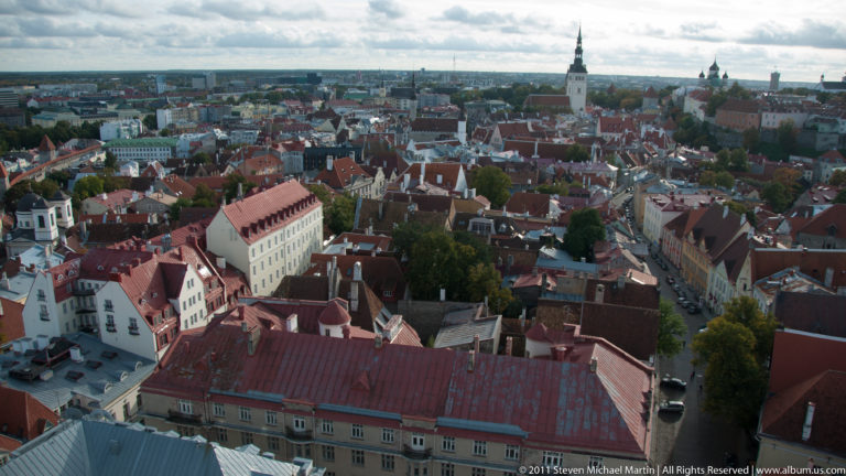 Elevated panorama of Tallinn Old Town with red-tiled roofs, narrow streets, historic buildings, and church towers across the skyline.
