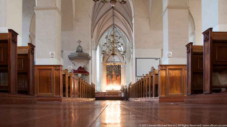 Low central view down a church nave with wooden pews on both sides, vaulted arches, chandeliers, and an illuminated altar.