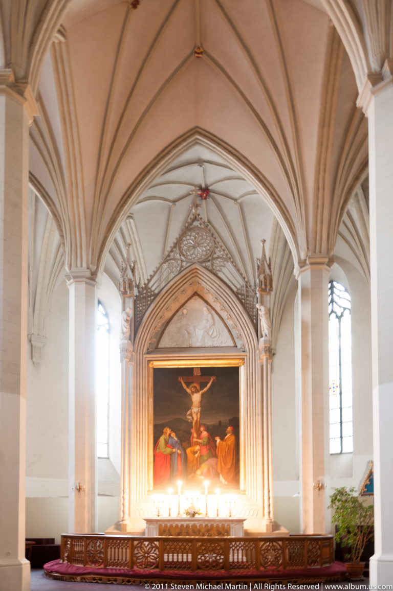 Crucifixion painting set in an ornate pointed altar frame beneath rib-vaulted ceilings inside a historic church.