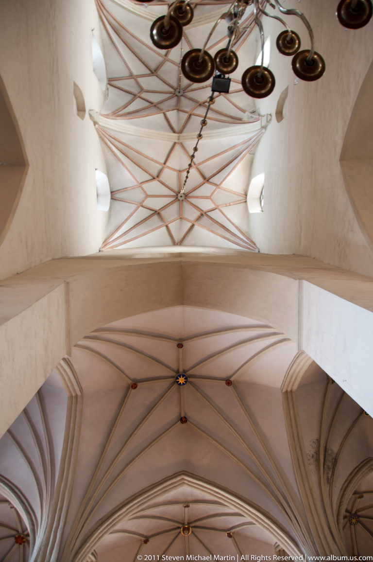 Upward view of rib-vaulted church ceilings with painted geometric lines and several suspended metal chandeliers.