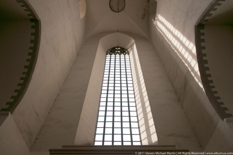 Very tall pointed church window with gridded glazing set into a high white plastered wall, lit by daylight.