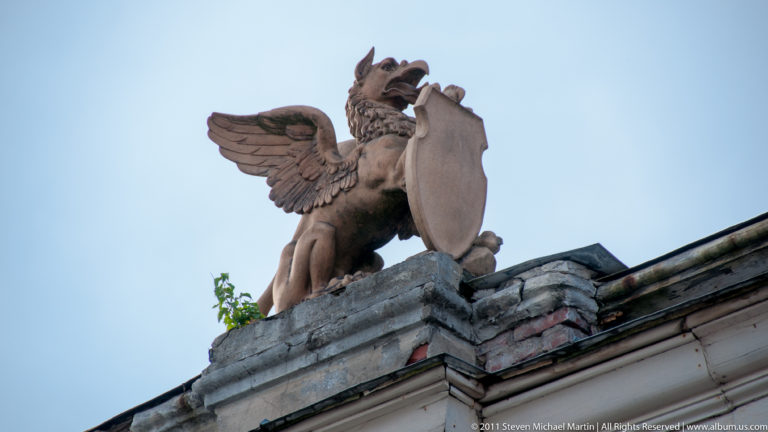 Winged griffin-like roof sculpture holding a shield on the edge of a historic building against a blue sky.