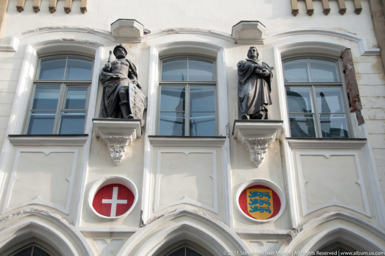 Two dark robed statues mounted on a white historic façade above red and yellow heraldic shields between tall windows.
