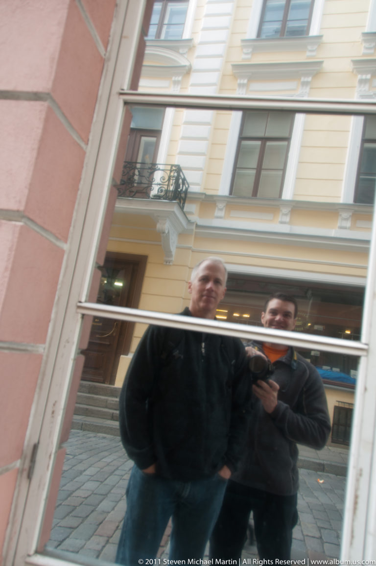 Reflection of two adults in a window, with pale historic façades and a Tallinn Old Town street visible behind them.