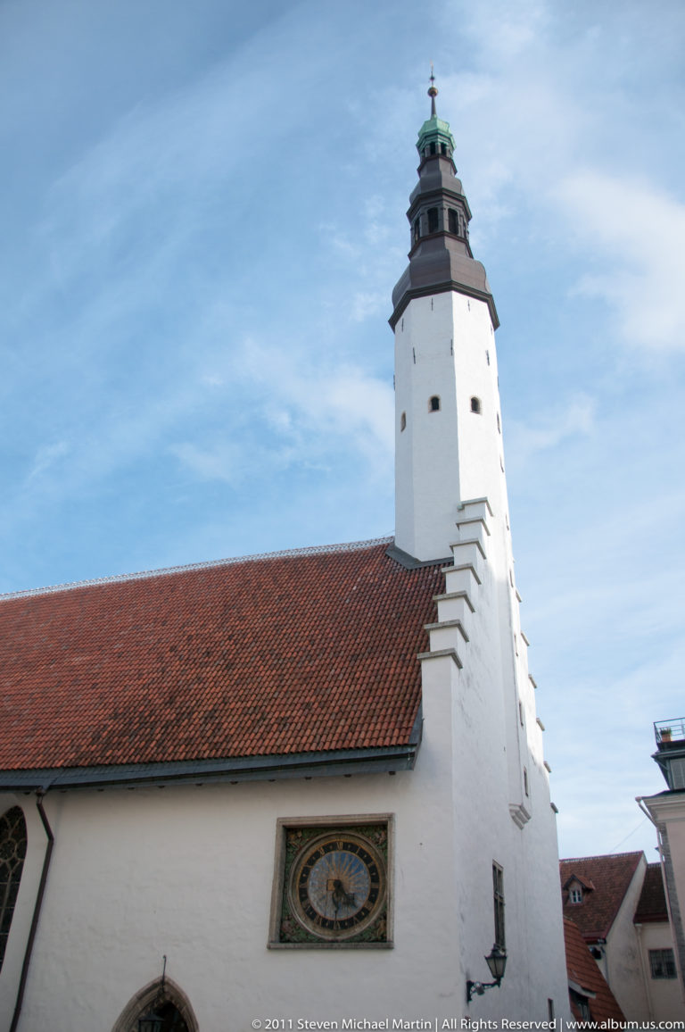 White church tower above a red-tiled roof in Tallinn Old Town, with a historic wall clock visible below the tower.