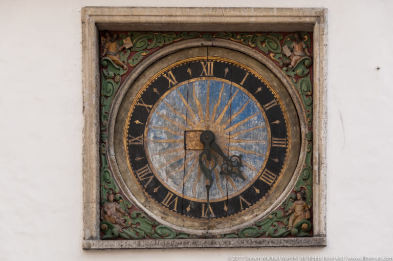 Ornate circular wall clock with Roman numerals, blue-and-gold dial, dark hands, and painted green decorative framing.