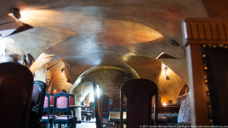 Warmly lit vaulted cellar interior with arched masonry bays, dining tables, chairs, and a narrow passage extending into the distance.