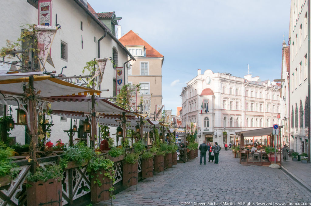 Pedestrian cobblestone street in Tallinn Old Town with planted restaurant terraces, historic façades, and people walking toward an intersection.