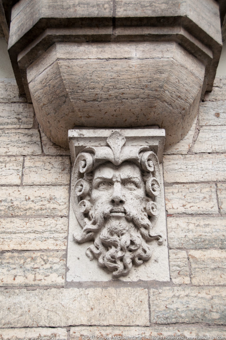 Carved grotesque stone face on a square block beneath a projecting masonry ledge.