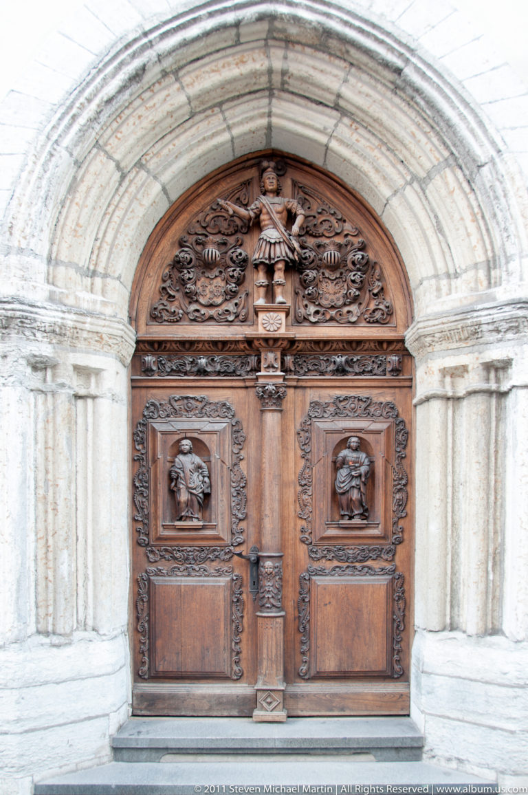 Ornately carved wooden double doors set within a rounded pale stone portal with decorative columns.