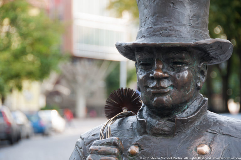 Bronze chimney sweep statue wearing a top hat and holding a round brush on a Tallinn street.