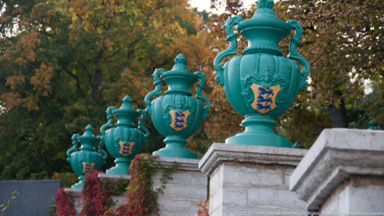 Row of turquoise ornamental urns on a stone balustrade, each bearing Estonia’s three-lions coat of arms.