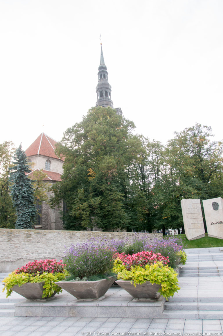 Colorful planters before the Eduard Vilde memorial, with trees and a historic tower in the background.