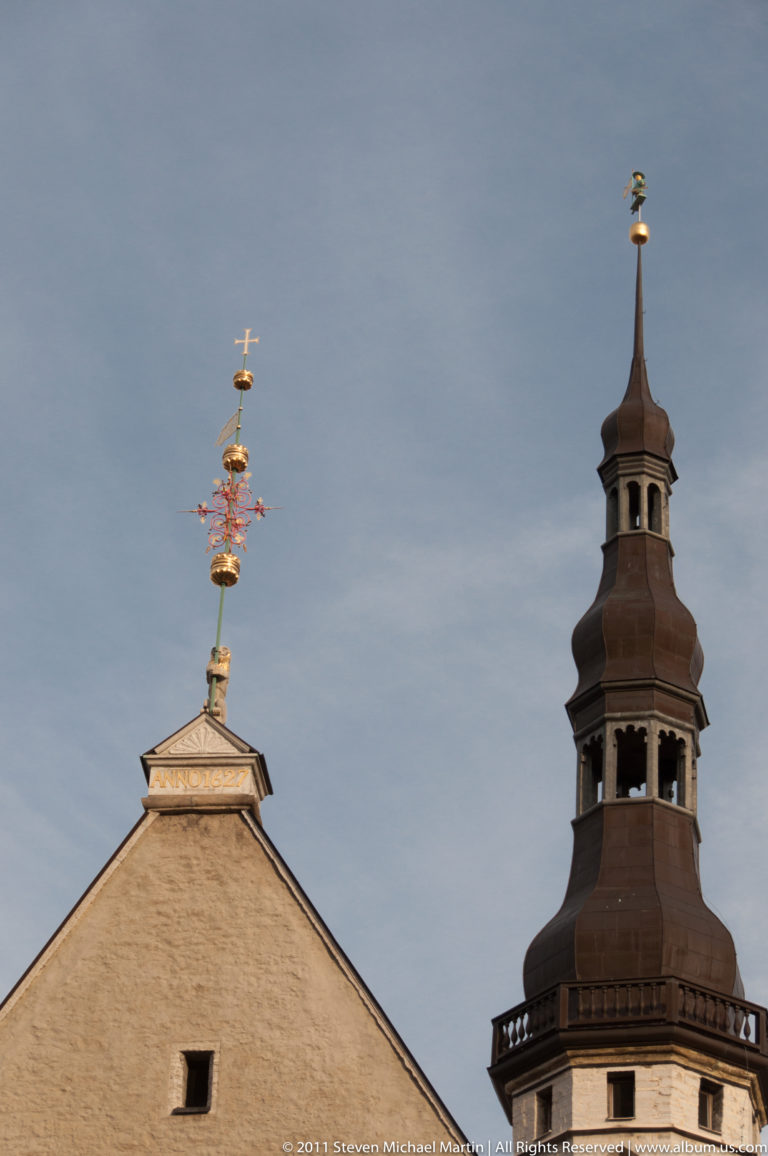 Historic Tallinn roofline with an elaborate metal finial beside a dark tiered tower.