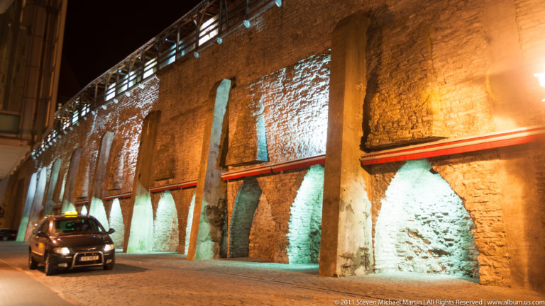 Long illuminated historic masonry wall with repeated recessed arches beside a narrow street at night.