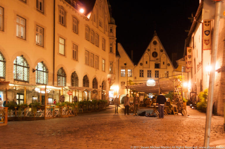 Cobbled Old Town street at night with café terraces, pedestrians, banners, and steep gabled façades.