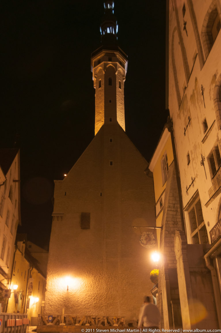 Tallinn Town Hall tower rising above a dark roof in a narrow Old Town night view.