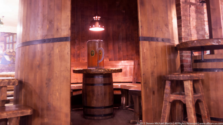 Wooden tavern interior with timber booths, barrel table, stools, and a beer mug under warm light.