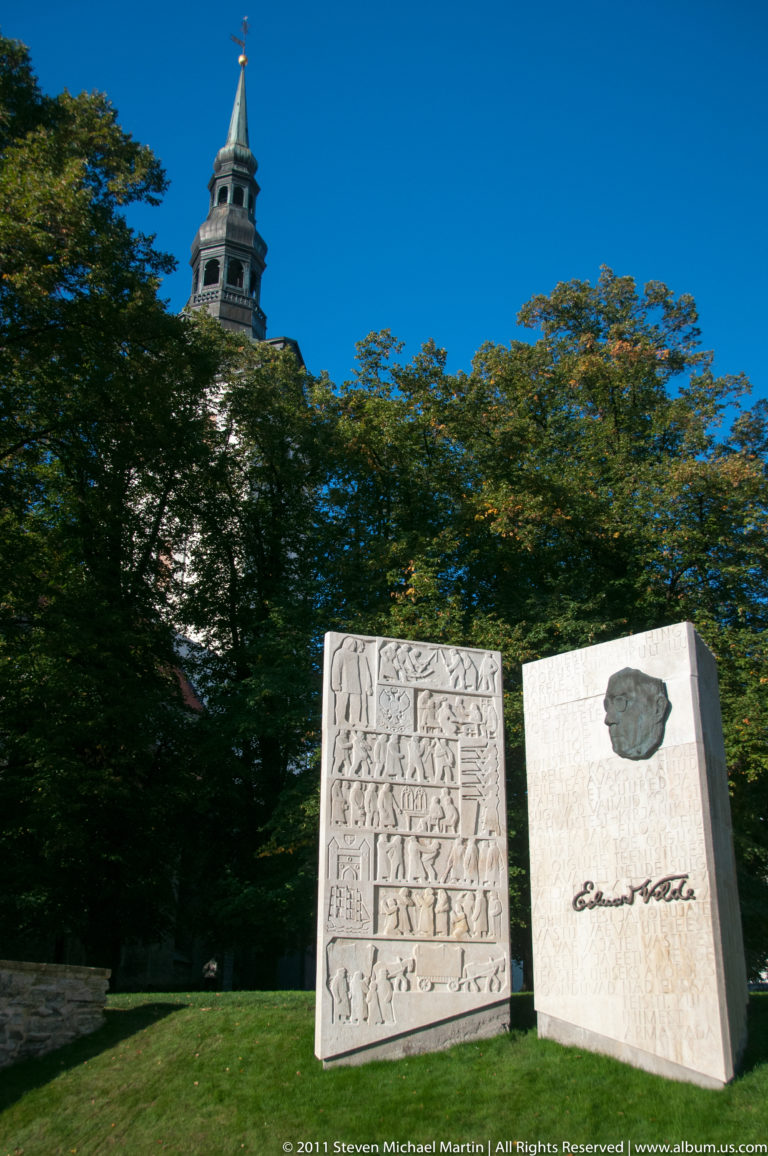 Eduard Vilde memorial on a lawn with trees and a historic tower rising behind it.