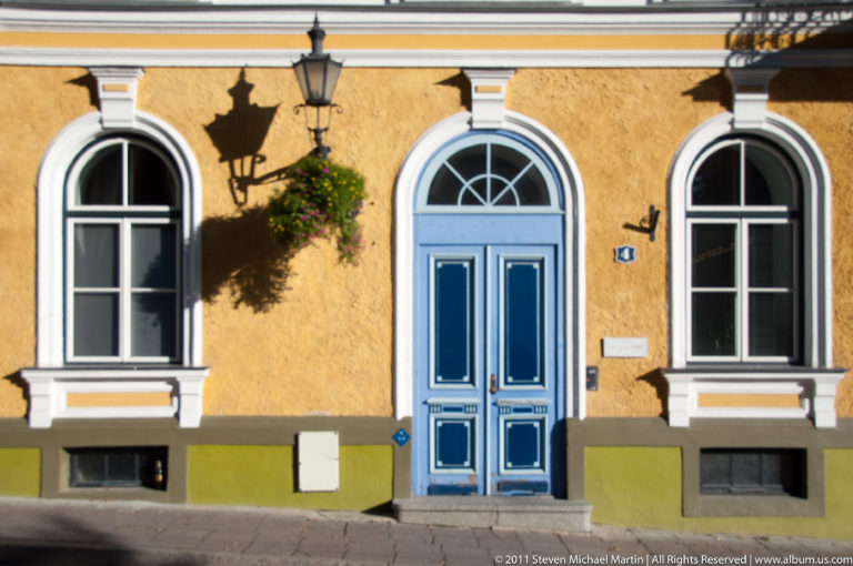 Blue paneled doorway between two arched windows on a sunlit ocher historic façade.