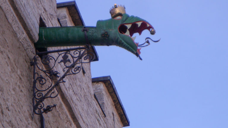 Green metal dragon rain spout with open red mouth projecting from a stone wall on an ornate iron bracket.