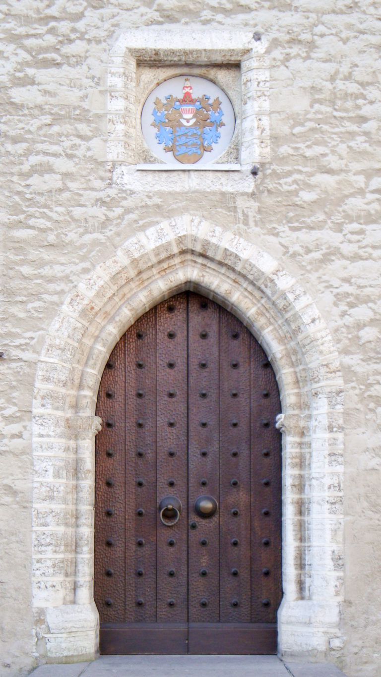 Dark studded wooden double doors with ring handles beneath a rounded stone arch and small heraldic plaque.