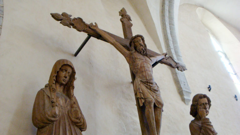 Wooden crucifix with carved Christ and mourning figures standing against a pale vaulted church wall.