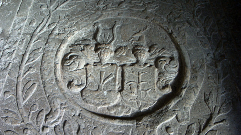 Carved heraldic shield with crests and scrolling foliage on a worn gray stone memorial slab.