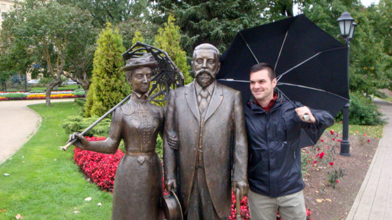 Man posing beside bronze statues of a woman and formally dressed man in a flower-lined Riga park.