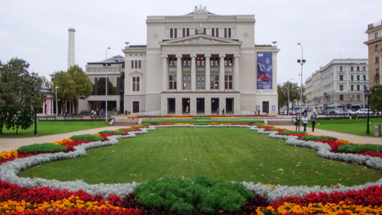 Latvian National Opera with columned Neoclassical façade beyond a lawn bordered by colorful formal flower beds.