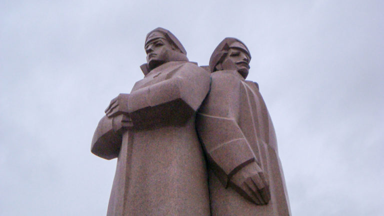 Low-angle view of two monumental stone figures in heavy cloaks standing back-to-back against the sky.