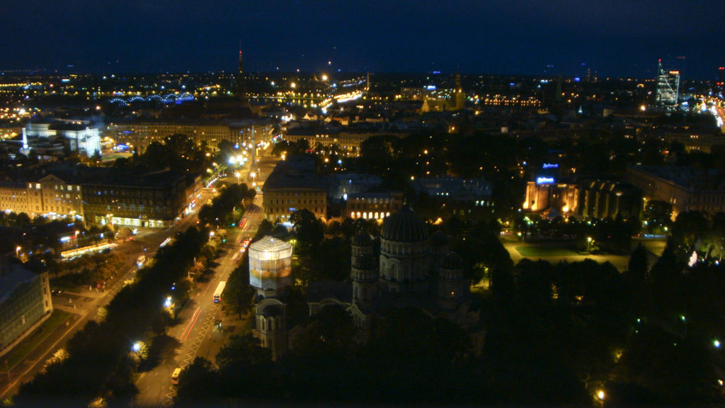 Elevated nighttime panorama of Riga with illuminated streets, dark parkland, church silhouettes, and distant city lights.