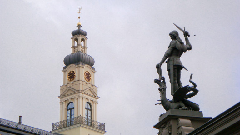 Dark warrior-and-dragon sculpture silhouetted beside a pale clock tower with an onion-shaped dome.