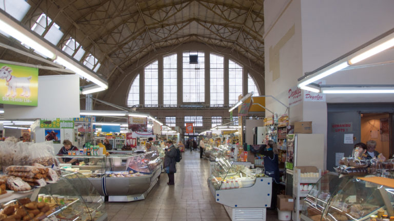 Long indoor market aisle with food counters and shoppers beneath a high steel roof and large arched window.